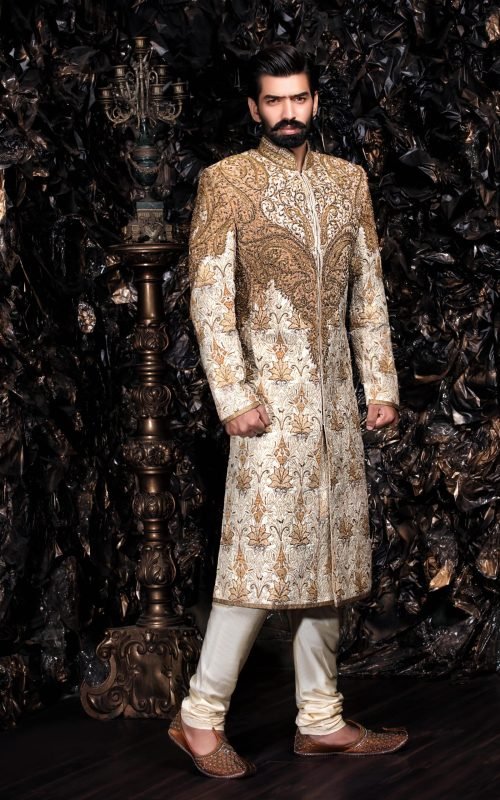 Man in ivory sherwani with intricate gold embroidery and mandarin collar posed against dramatic backdrop of antique brass decorative elements